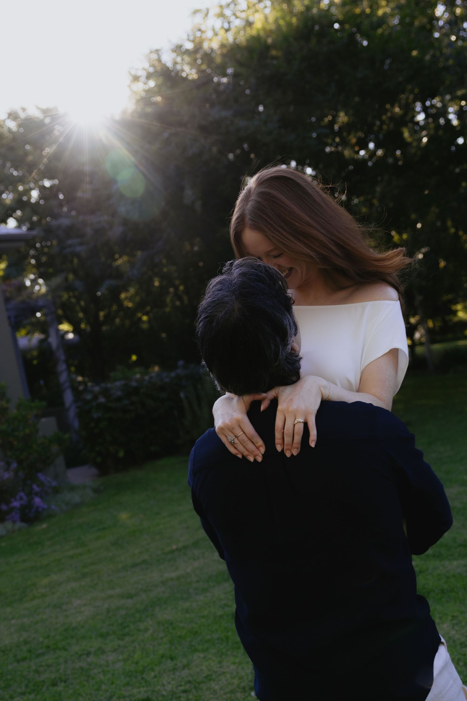 Couple embracing in a sunlit garden, woman in white off-shoulder top smiling, golden hour backlight.