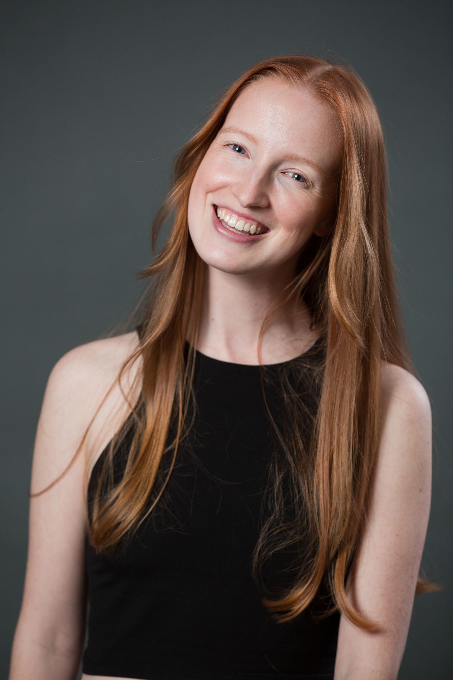 Smiling young woman with long red hair wearing a black sleeveless top against a gray background.