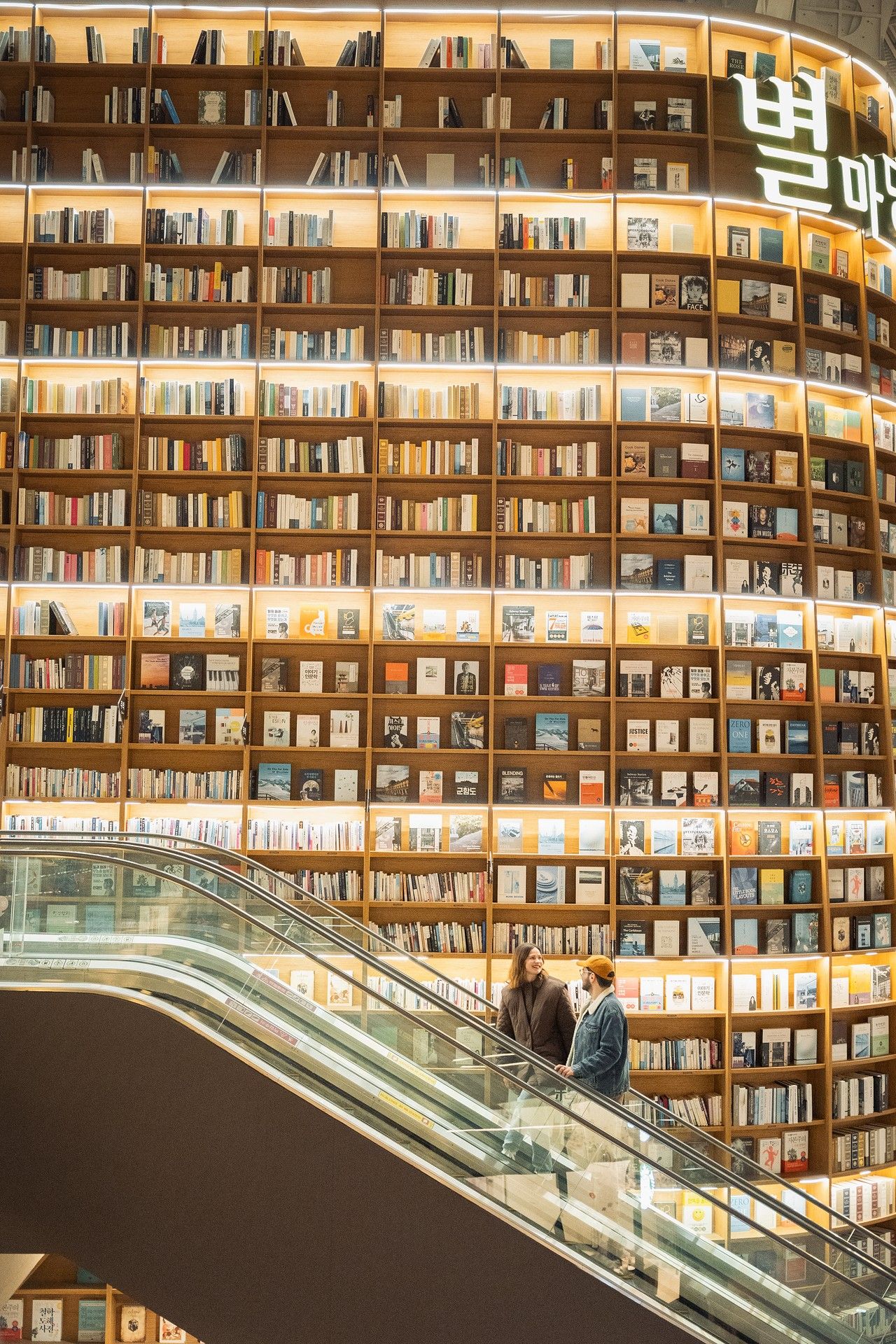 Two people ride an escalator beside a massive curved bookshelf wall filled with books in a modern Korean library.