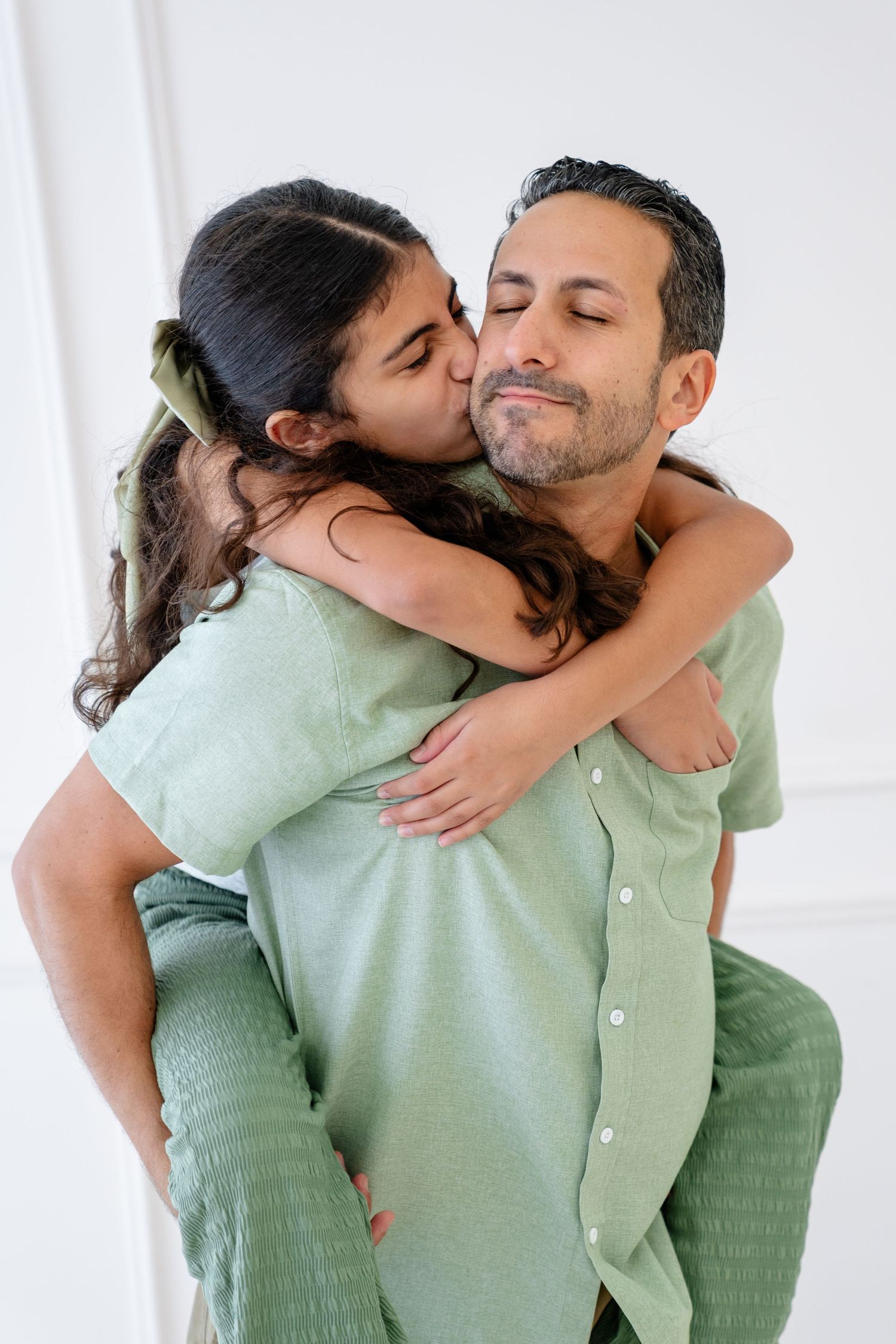 A young girl gives a piggyback ride kiss on the cheek to a smiling man in a green shirt indoors.