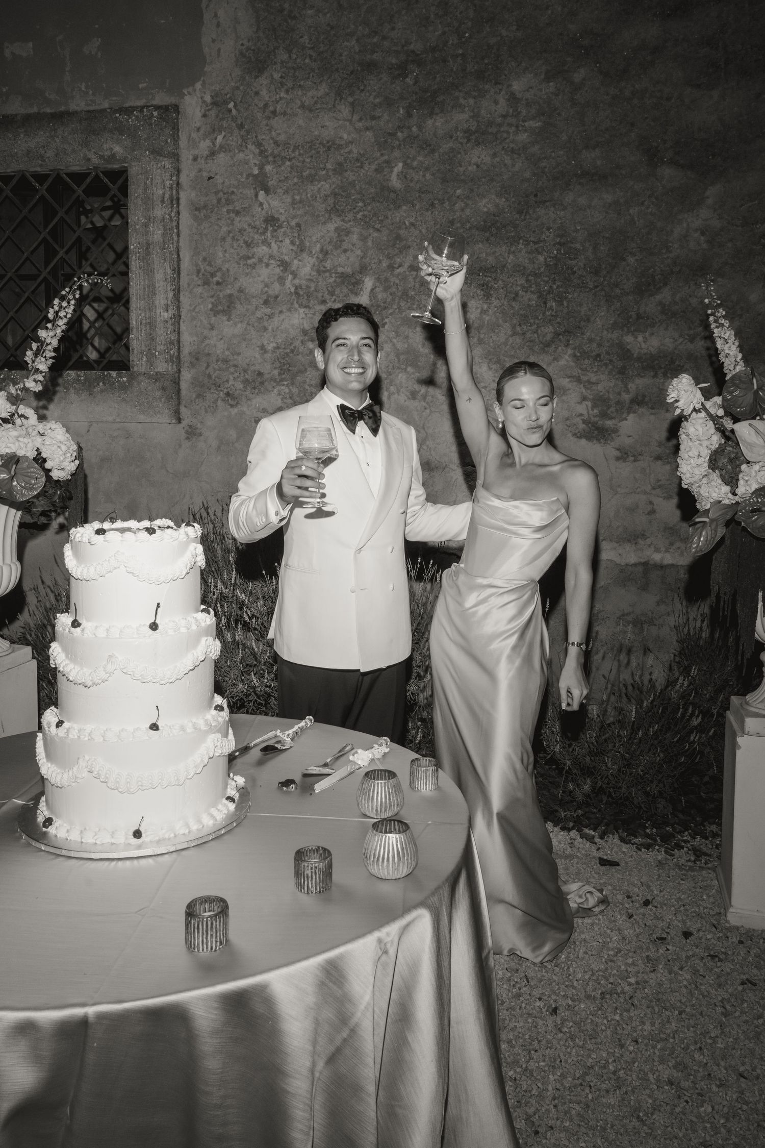 Black and white photo of a couple toasting with drinks beside a tiered wedding cake at an elegant outdoor reception.