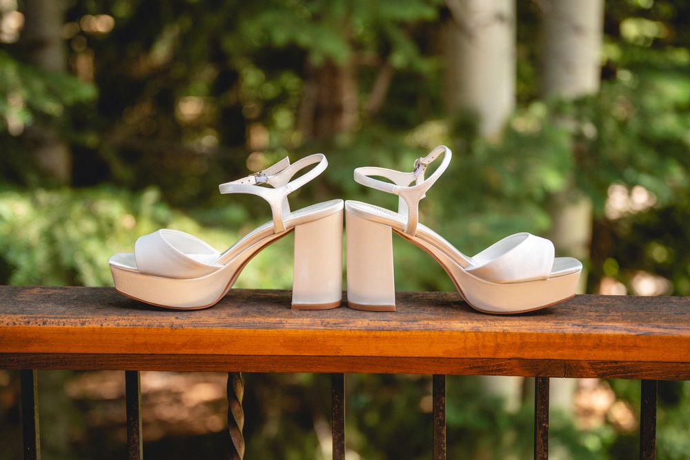 White platform block heel bridal sandals displayed on a wooden railing with lush green trees in the background.
