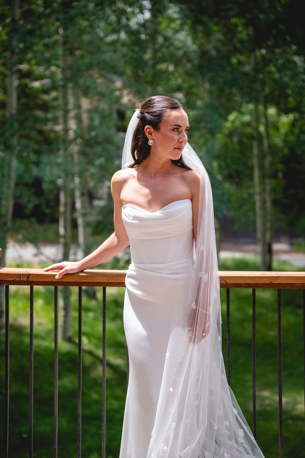 Bride in strapless white gown and veil stands on outdoor balcony surrounded by lush green trees.