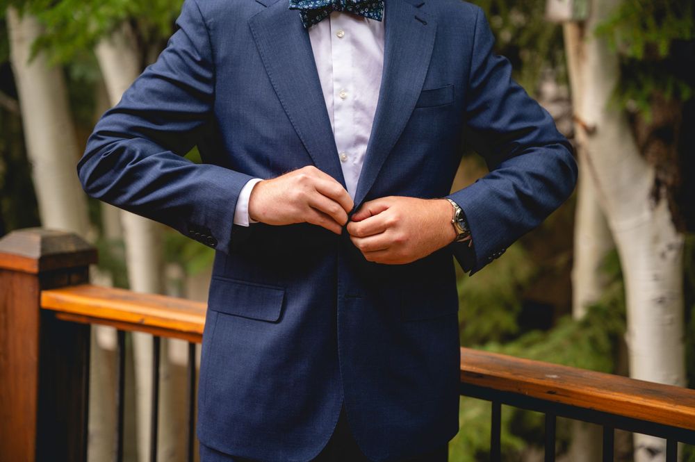 Man in navy blue suit buttoning jacket with colorful bow tie and watch, standing on wooden deck outdoors.