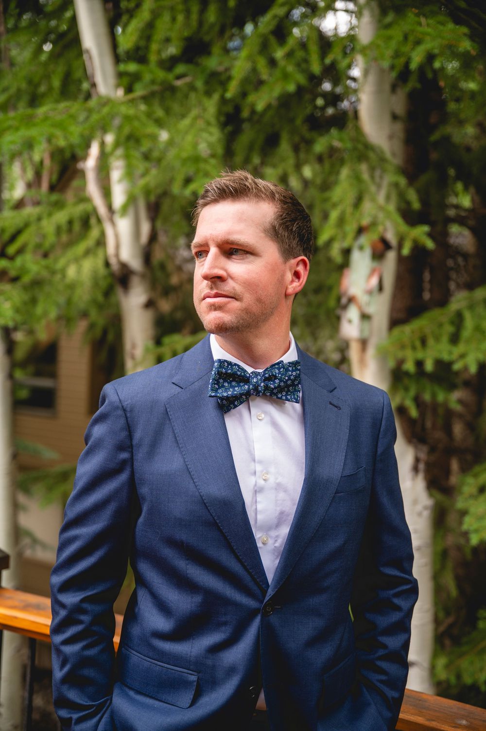 A groom in a navy blue suit and floral bow tie stands outdoors among trees.