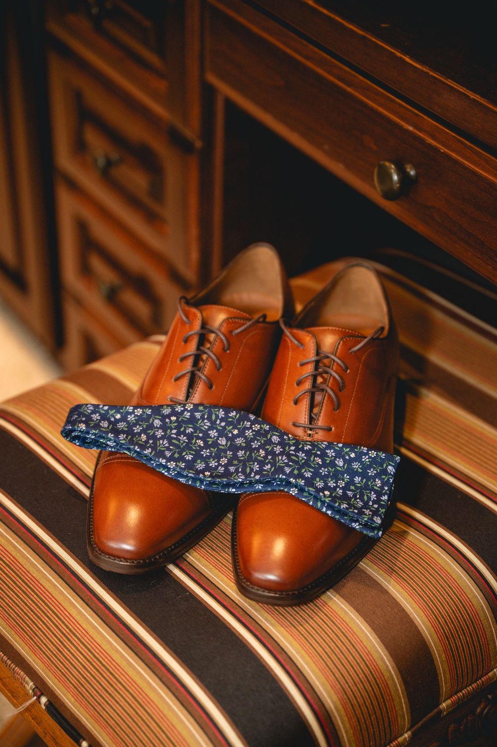 Brown leather Oxford shoes with a navy floral bow tie resting on a striped surface near wooden drawers.