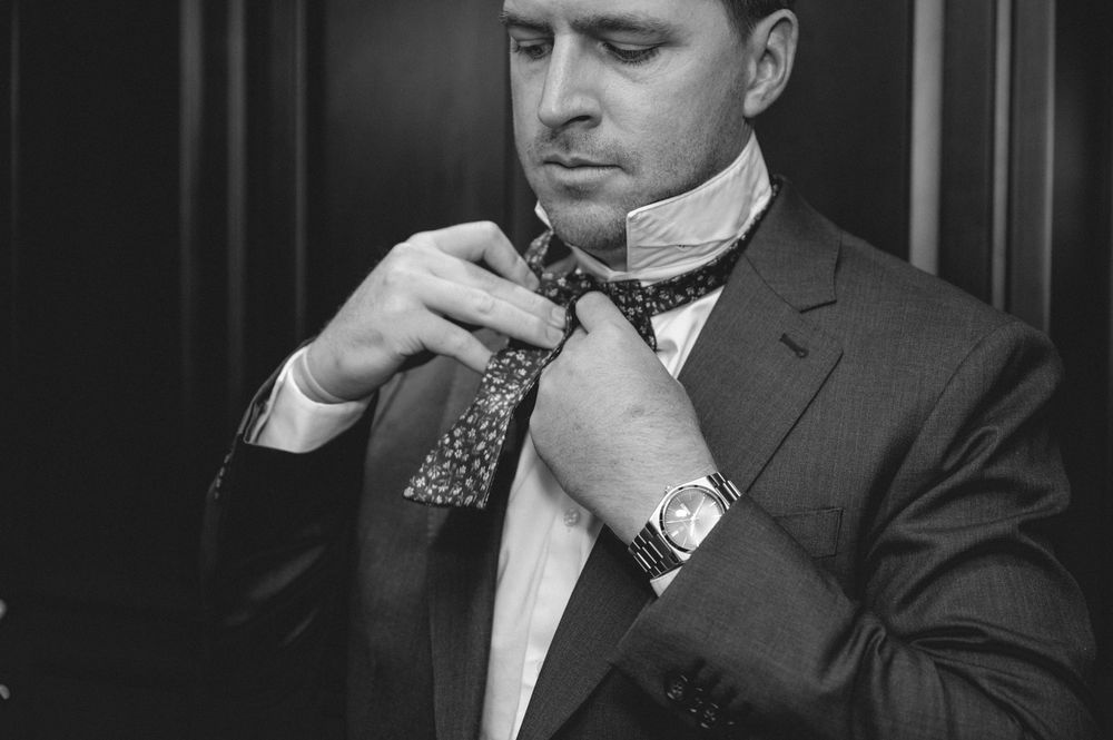 Black and white photo of a man in a suit adjusting his bow tie, wearing a watch, elegant formal attire.
