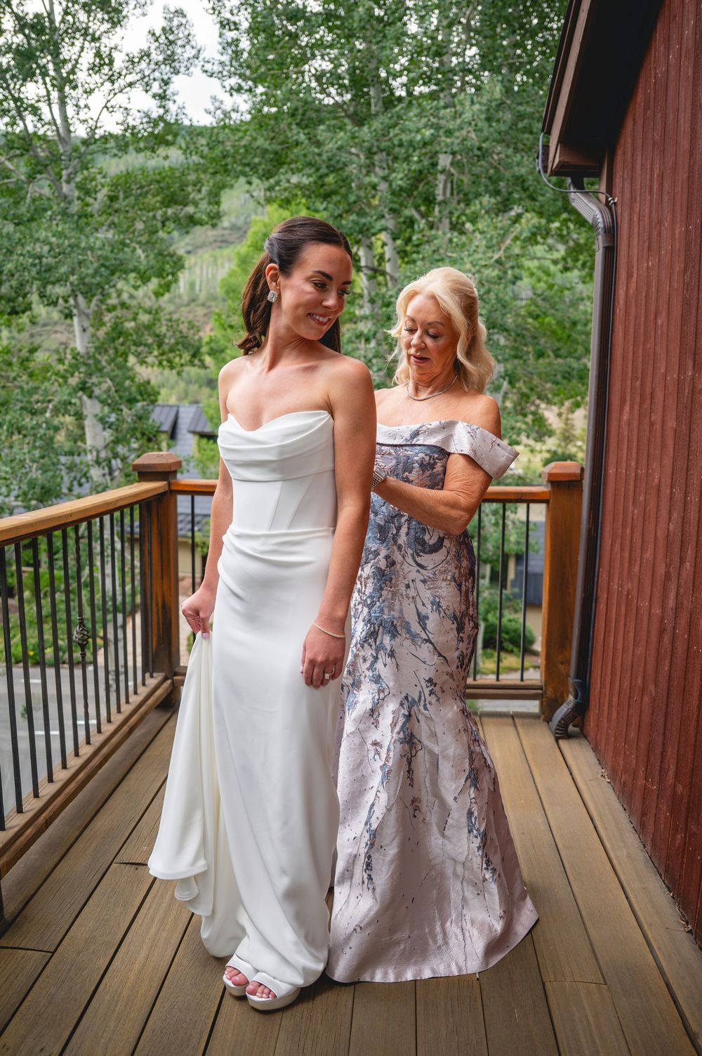 Bride in white strapless gown with older woman in floral dress on wooden deck surrounded by trees.