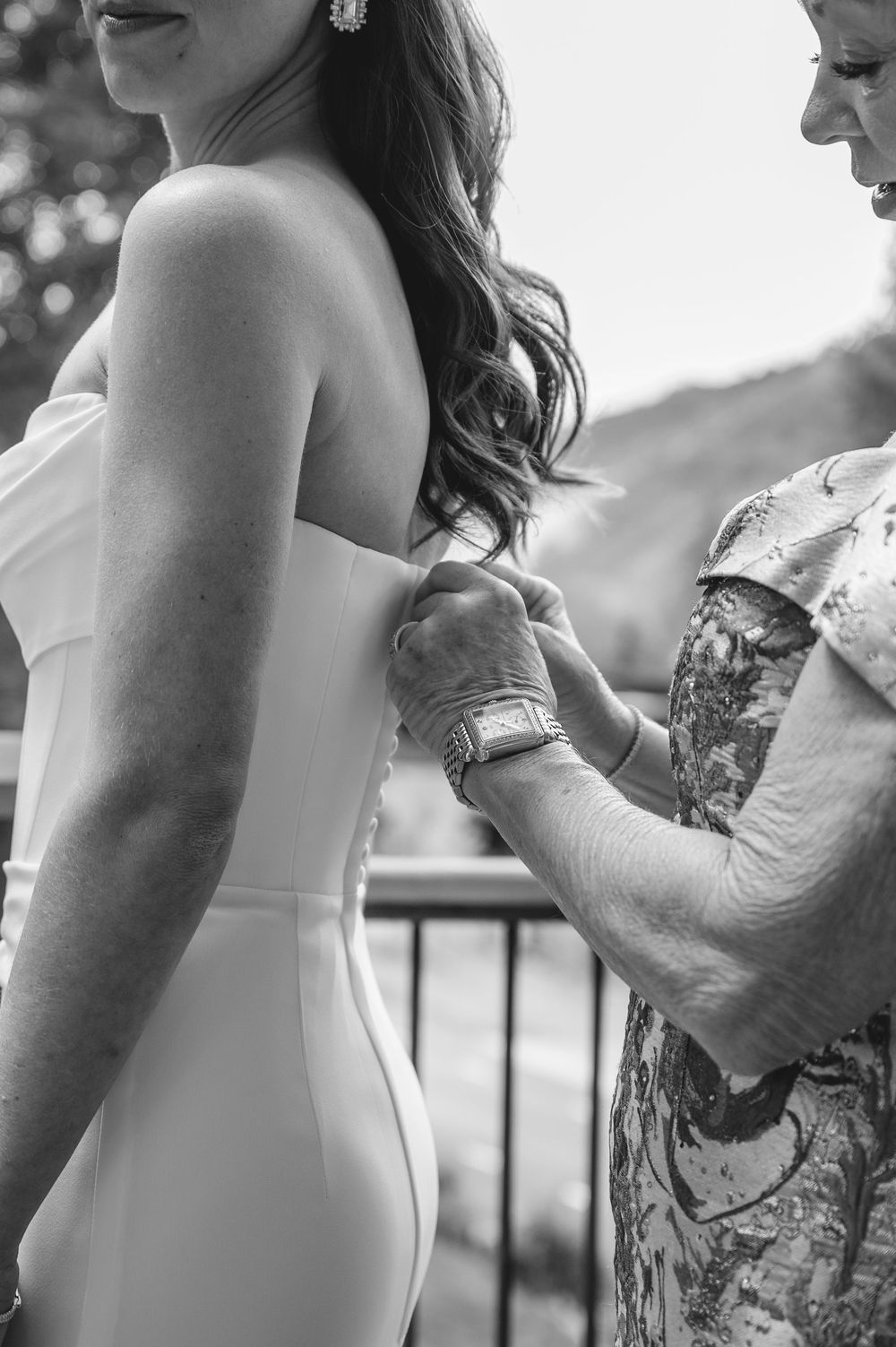 Black and white photo of a bride in a strapless gown being helped by an older woman on a balcony.