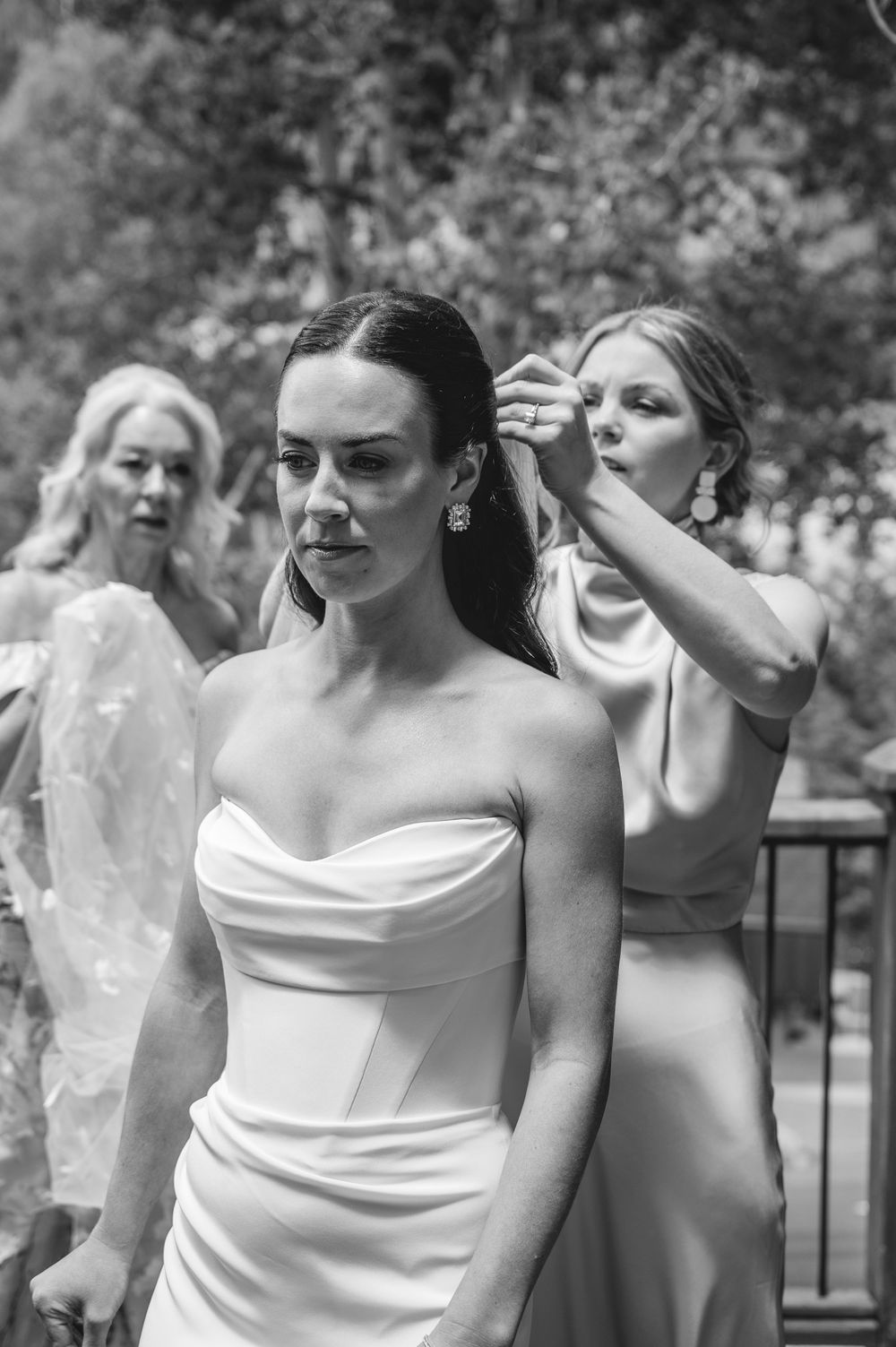 Bride in strapless gown gets hair adjusted by bridesmaid outdoors, with another woman in background.