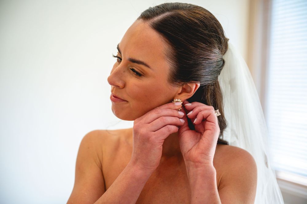 Bride in a white veil putting on gold earrings while getting ready for her wedding day.