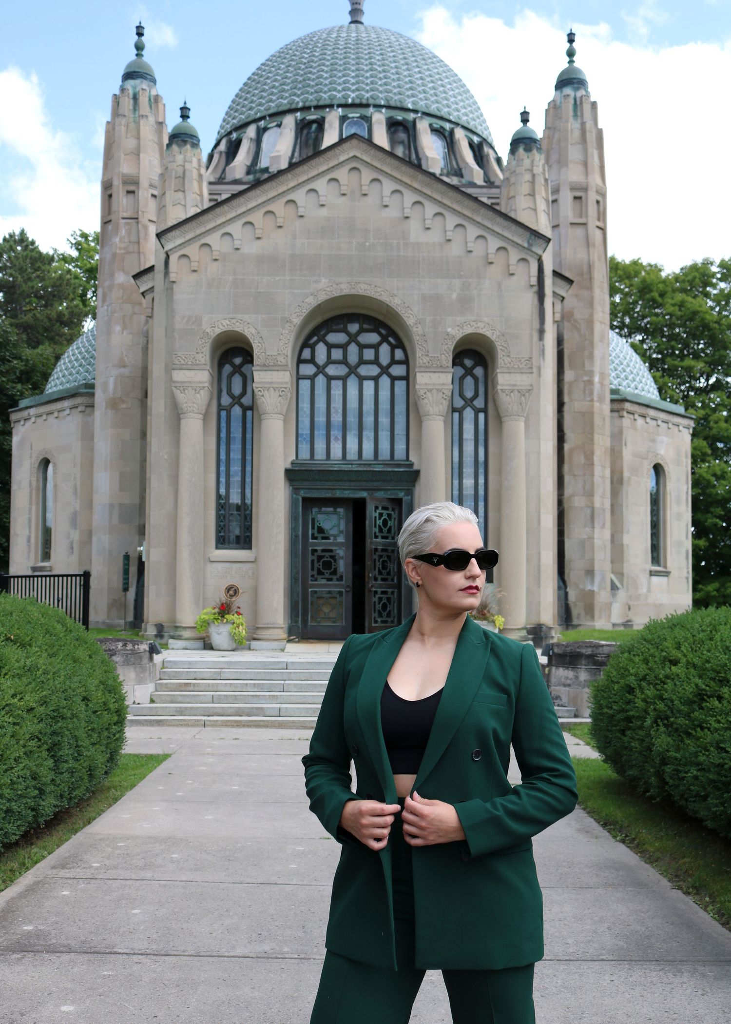 Woman in green suit poses before ornate domed stone building with arched windows and decorative spires.