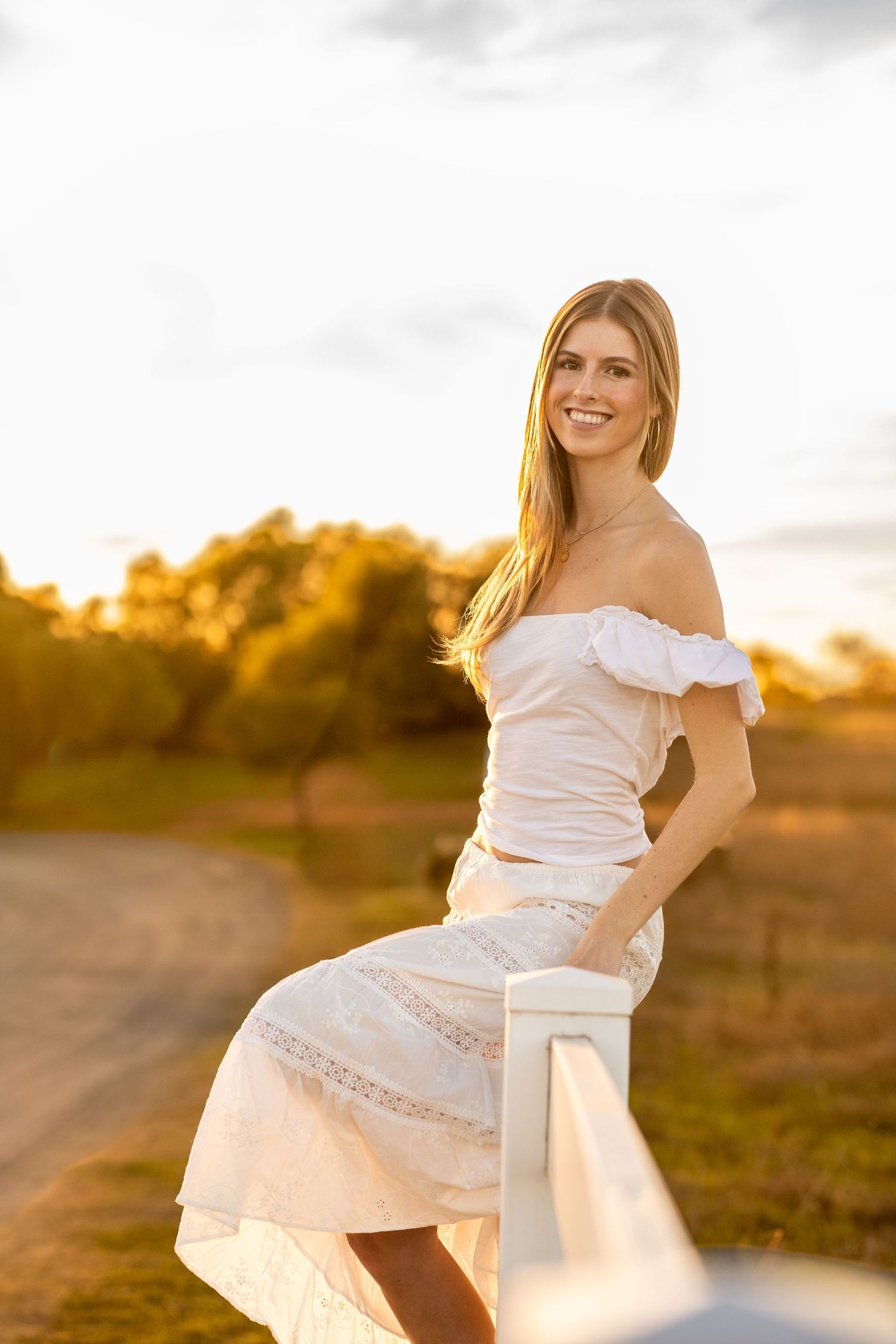 Young woman in white off-shoulder dress smiling on a rural road at golden sunset hour.