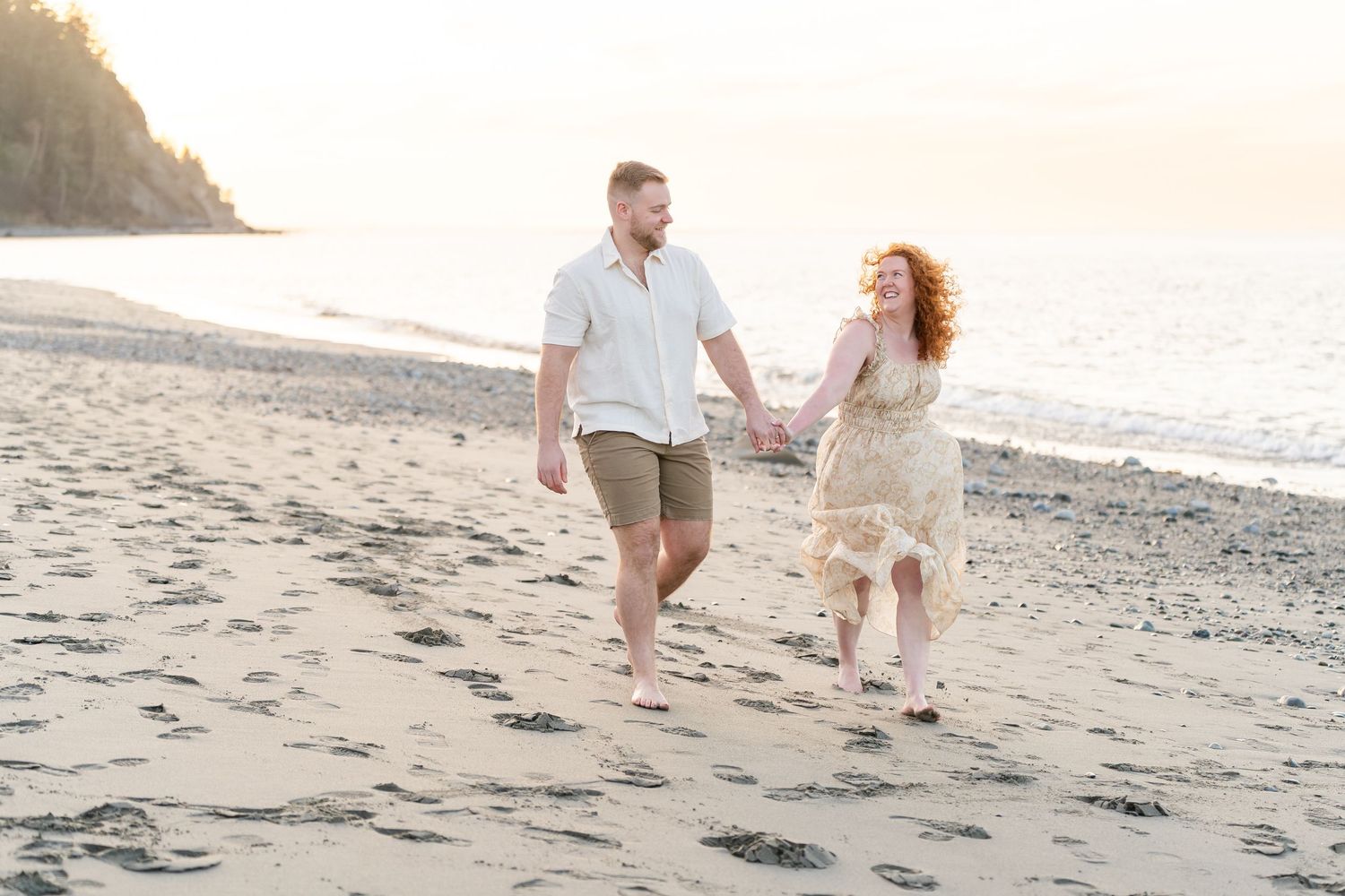 Couple holding hands walking barefoot on sandy beach at golden hour sunset with ocean waves in background.