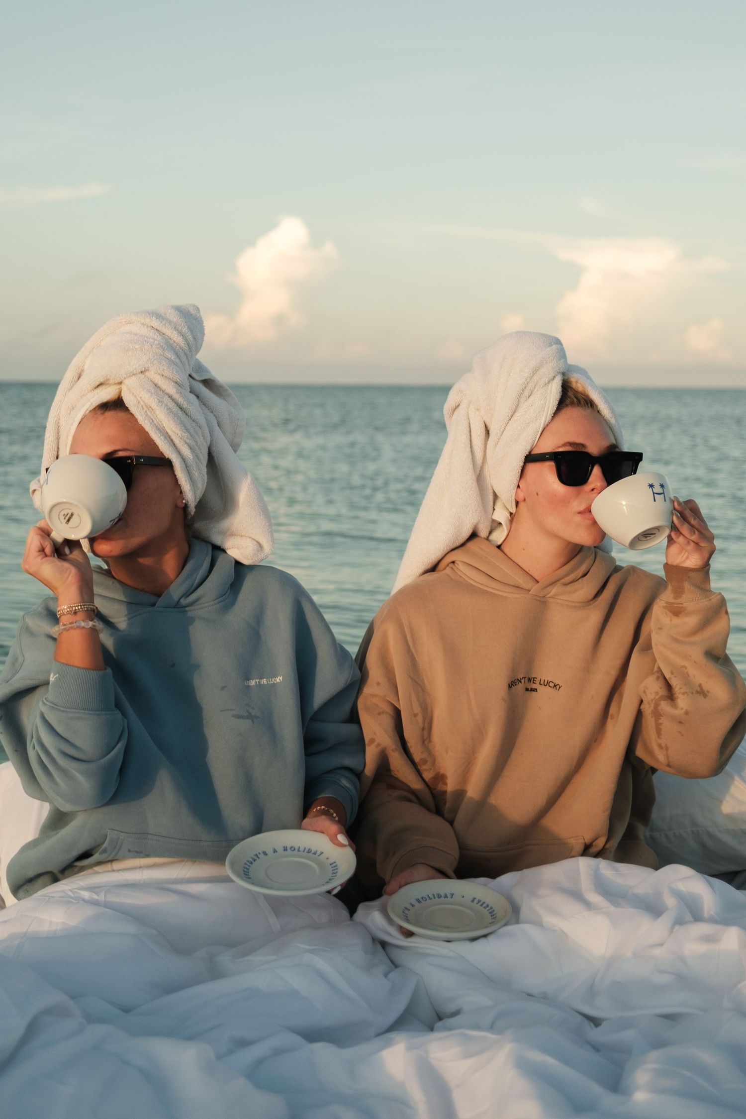 Two women with towels on heads sip coffee by the ocean in matching hoodies, enjoying a relaxing beach morning.