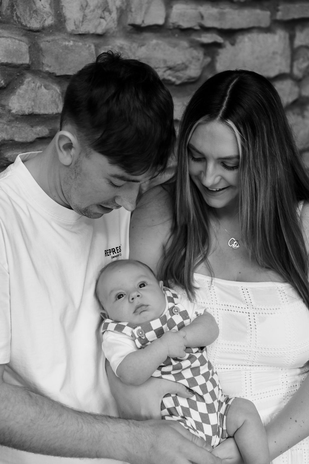 Black and white photo of a young couple smiling at their newborn baby, posed against a stone wall backdrop.