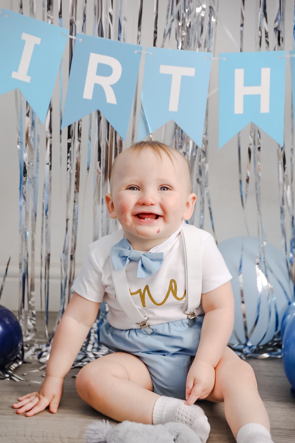 Smiling baby boy in blue outfit celebrating first birthday with colorful balloons and birthday banner backdrop.