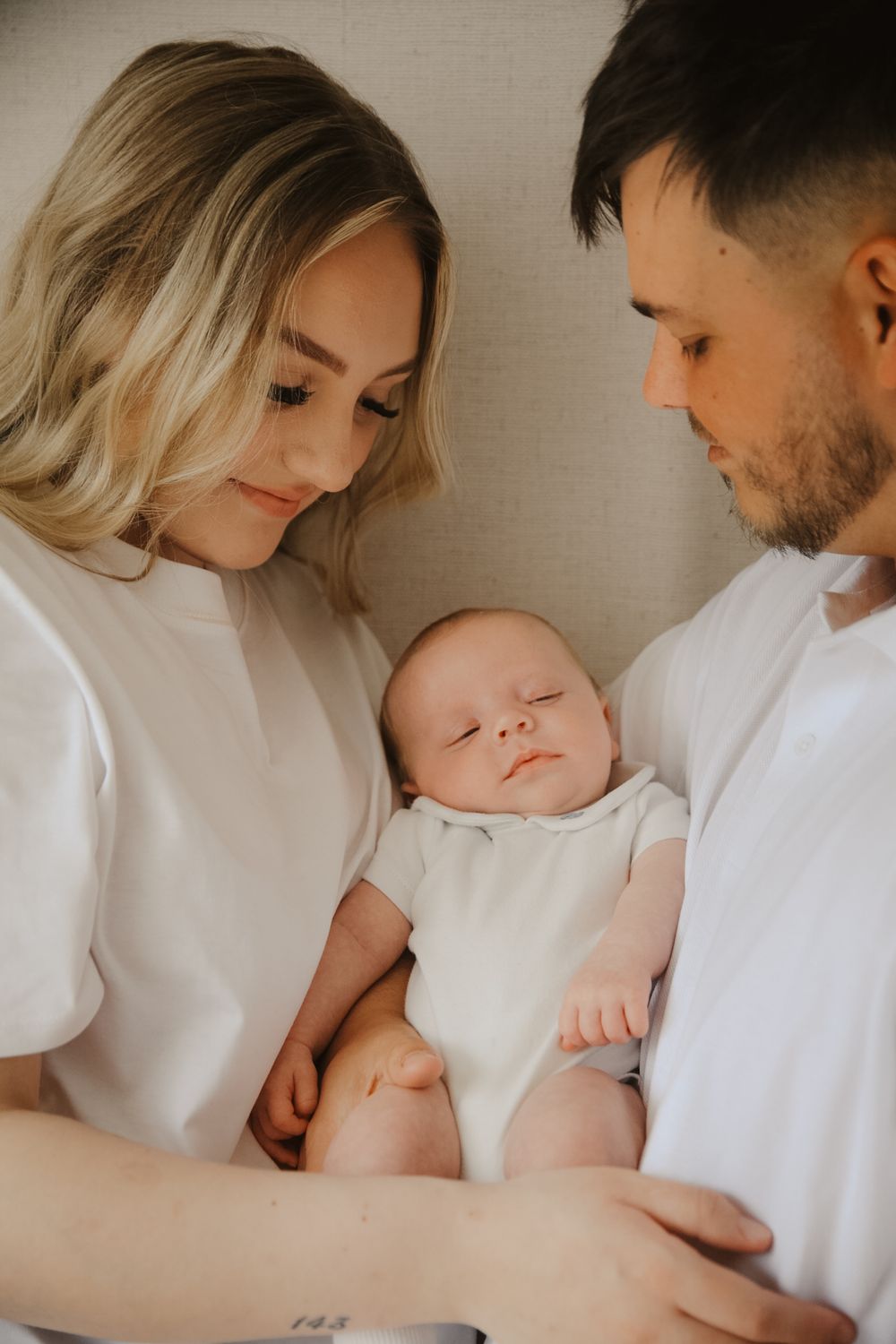 Young parents in white shirts tenderly holding their sleeping newborn baby in a cozy home setting.