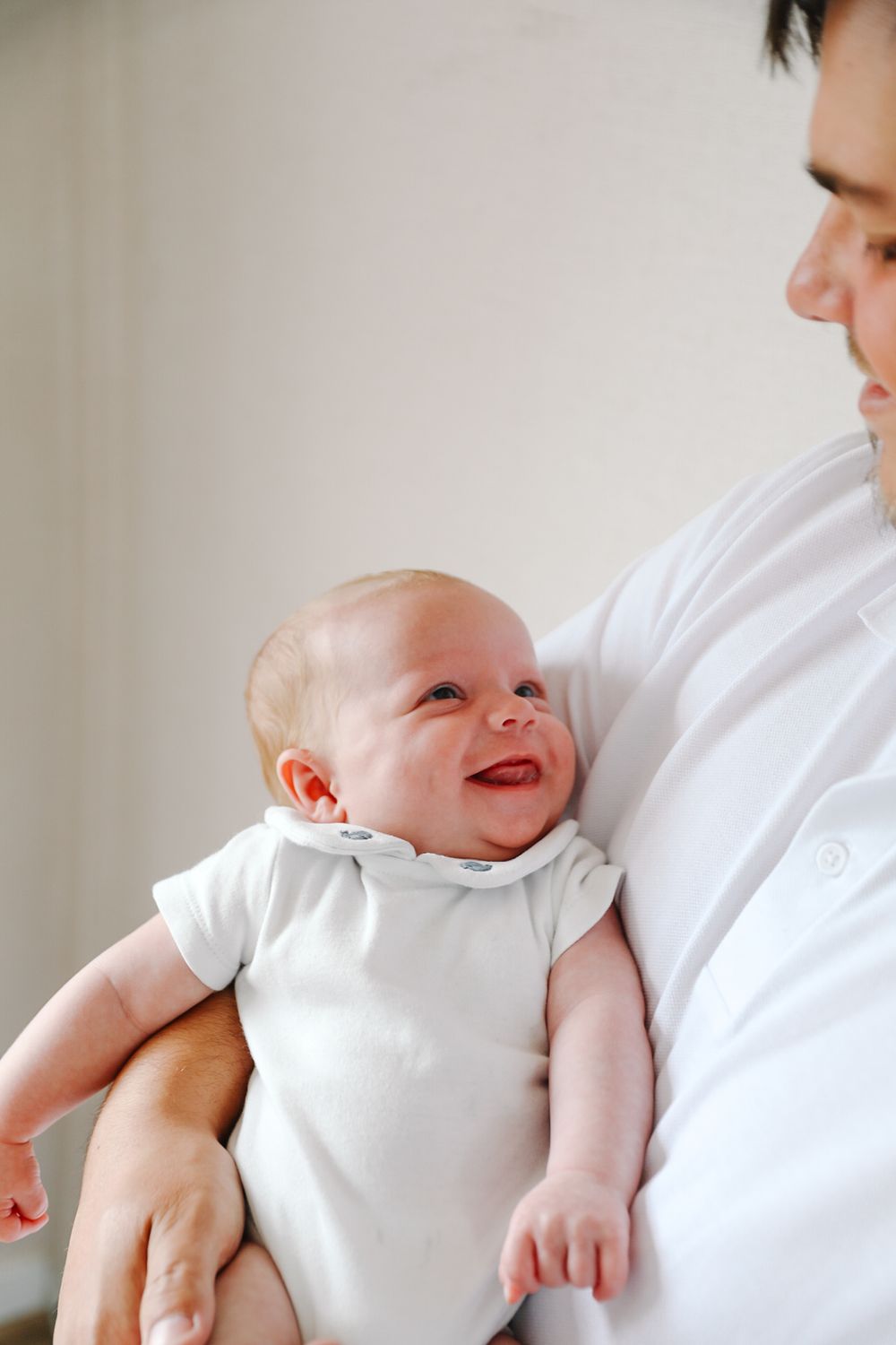 A smiling newborn baby in a white onesie being cradled in the arms of an adult wearing a white shirt.