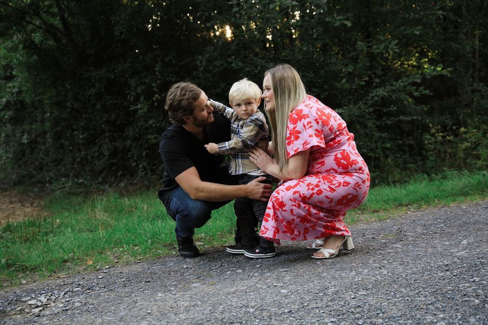Family of three posing outdoors on gravel path, parents crouching beside toddler with lush green trees behind.