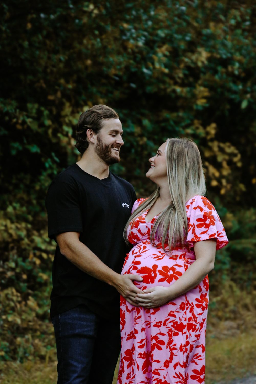 Smiling couple cradles pregnant belly outdoors, woman in red floral dress, man in black shirt, lush green foliage background.
