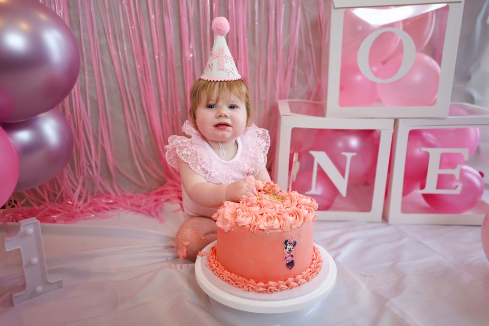 Baby girl in pink outfit and party hat sits beside a pink Minnie Mouse smash cake with ONE balloon boxes.