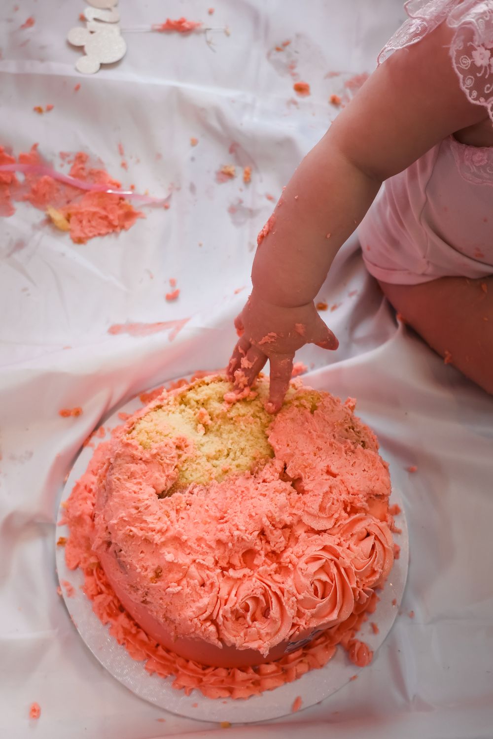 Baby smashing pink frosted first birthday cake, with frosting scattered on white cloth during cake smash celebration.