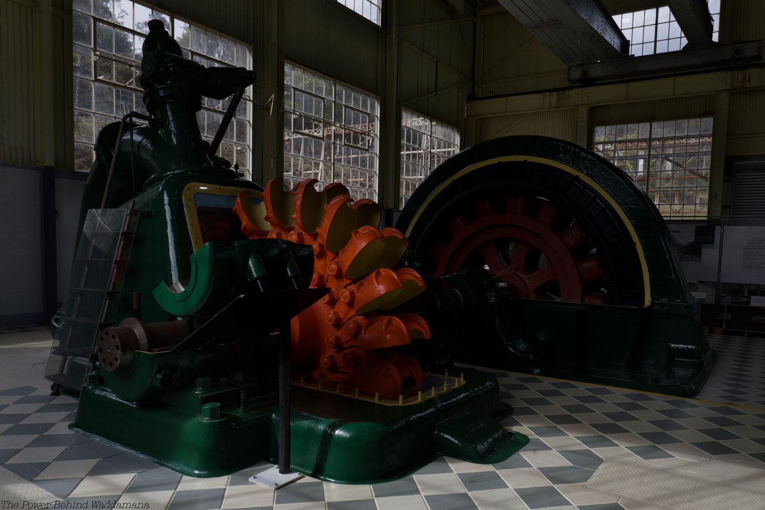 Vintage green industrial generator with orange turbine blades inside a historic power plant hall with large windows and checkered tile floor