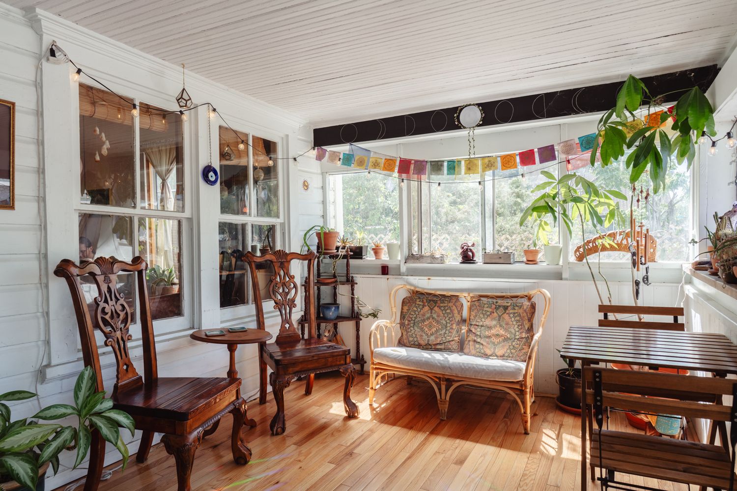 Bright sunroom with wood floors, carved wooden chairs, a cushioned bench, hanging plants, a colorful bunting garland and tall windows.