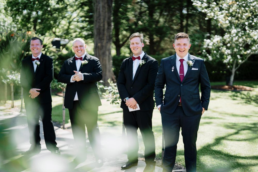 Four groomsmen in black suits and bow ties stand together outdoors in a sunlit garden, smiling before a wedding ceremony.