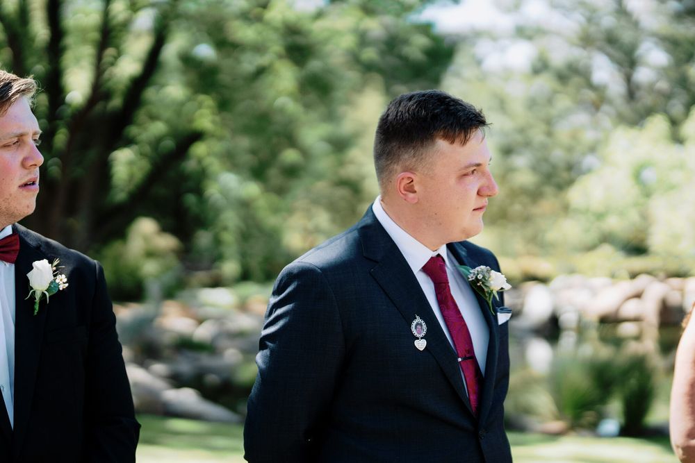 Two groomsmen in dark suits with white boutonnieres stand outdoors at a wedding ceremony, surrounded by green trees and soft sunlight.