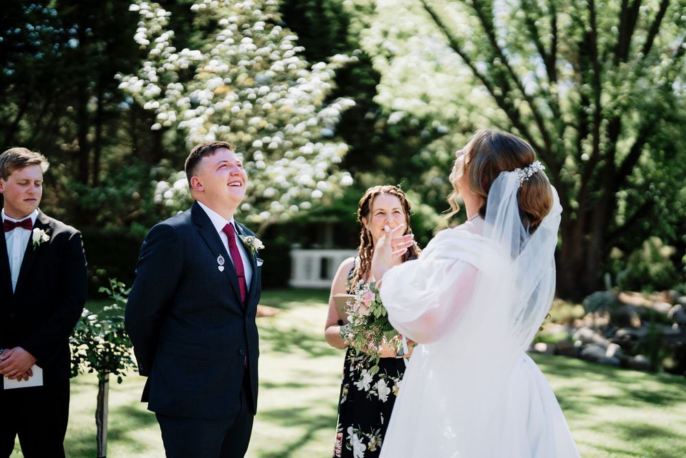 Bride in a white dress and veil raises her hand to her face during an outdoor garden wedding ceremony as the groom and a bridesmaid look on.