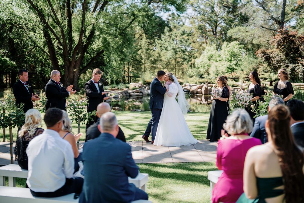 Bride and groom embrace in an outdoor garden wedding ceremony as seated guests and wedding party look on beneath tall shady trees.