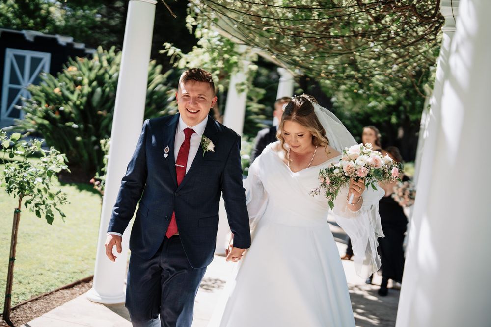 Smiling bride in a white gown holding a pink floral bouquet walks hand in hand with a groom in a navy suit and red tie outdoors.