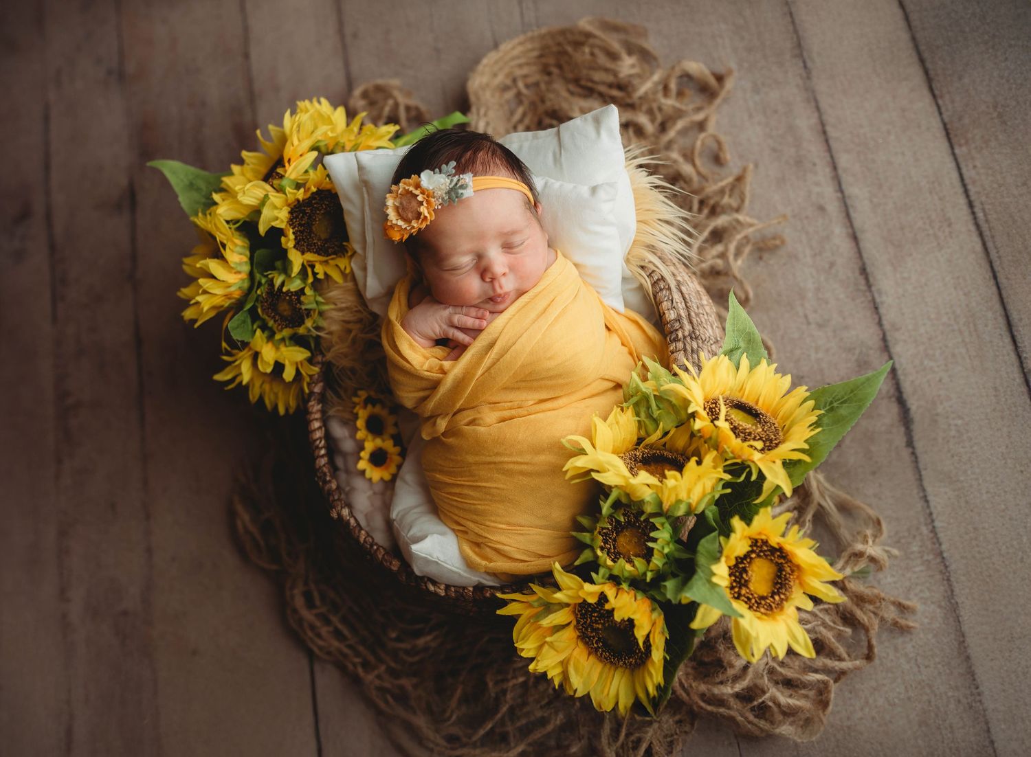 Sleeping newborn baby in a yellow wrap and floral headband resting in a woven basket surrounded by sunflowers on a rustic wooden floor.
