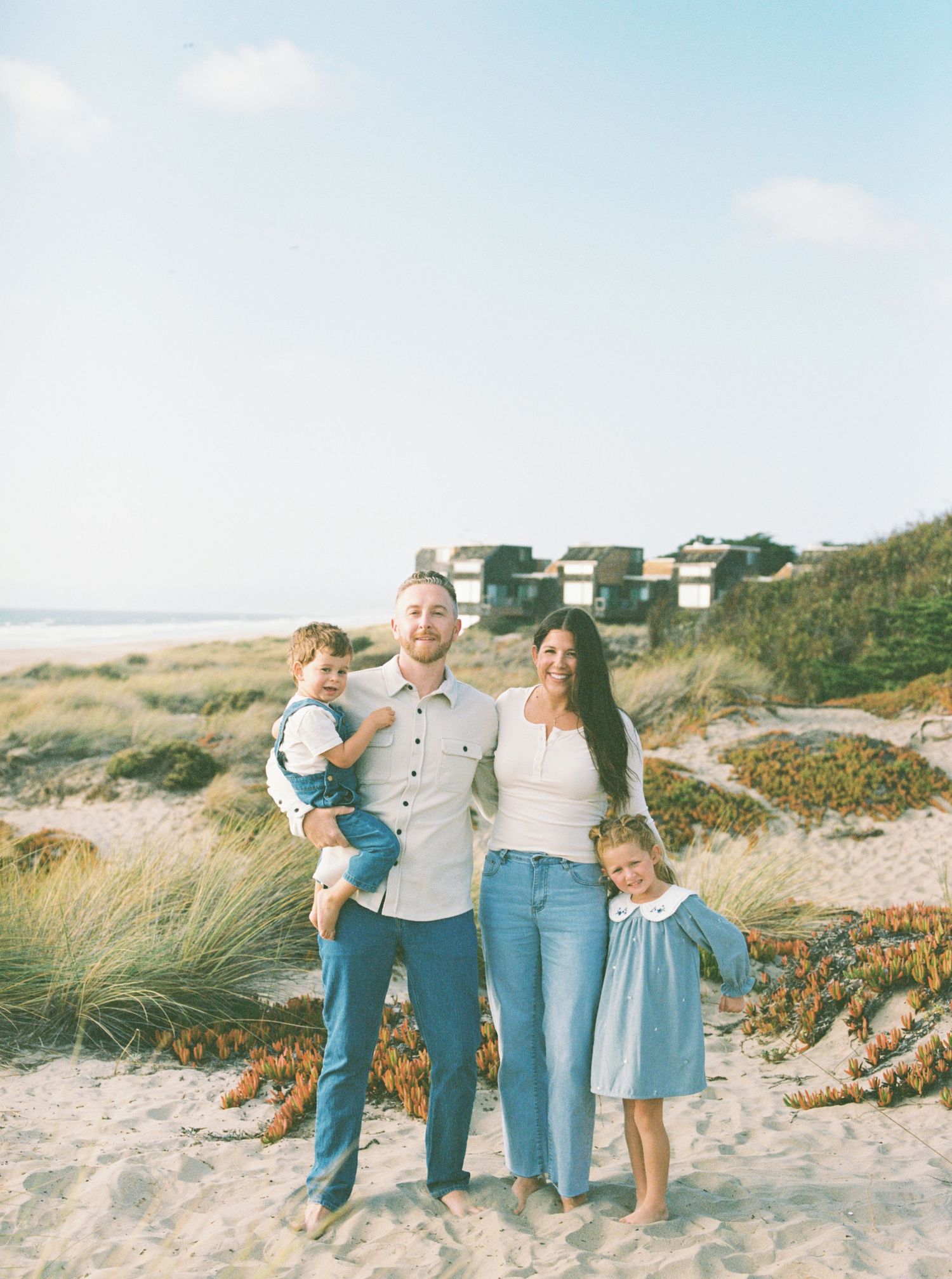 Smiling family of four in matching blue and white outfits posing together on a sandy beach with dune grass and coastal buildings behind.