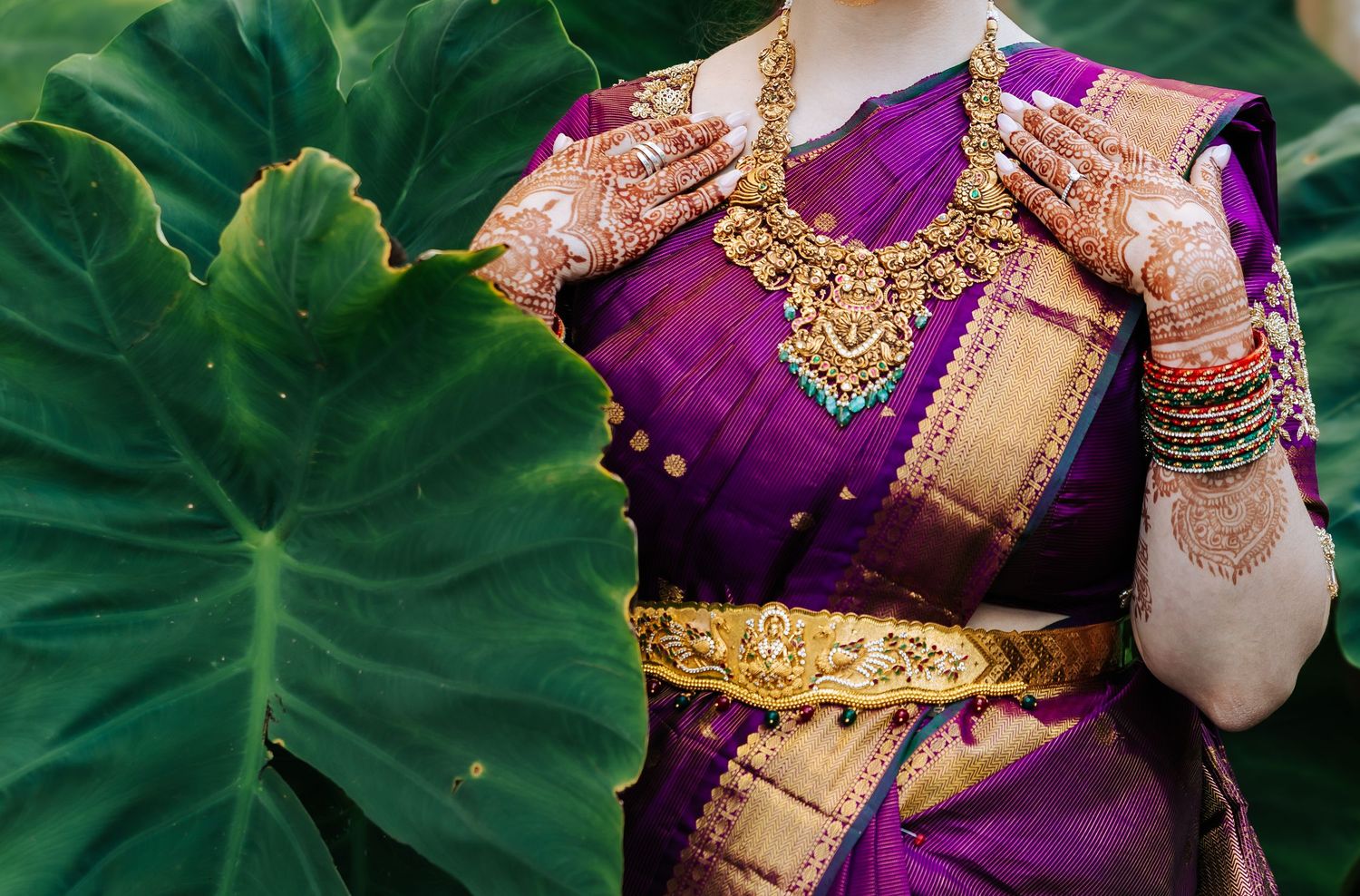 Bride in a purple and gold silk saree with temple jewelry, gold waist belt, bangles and henna-decorated hands beside large green leaves.