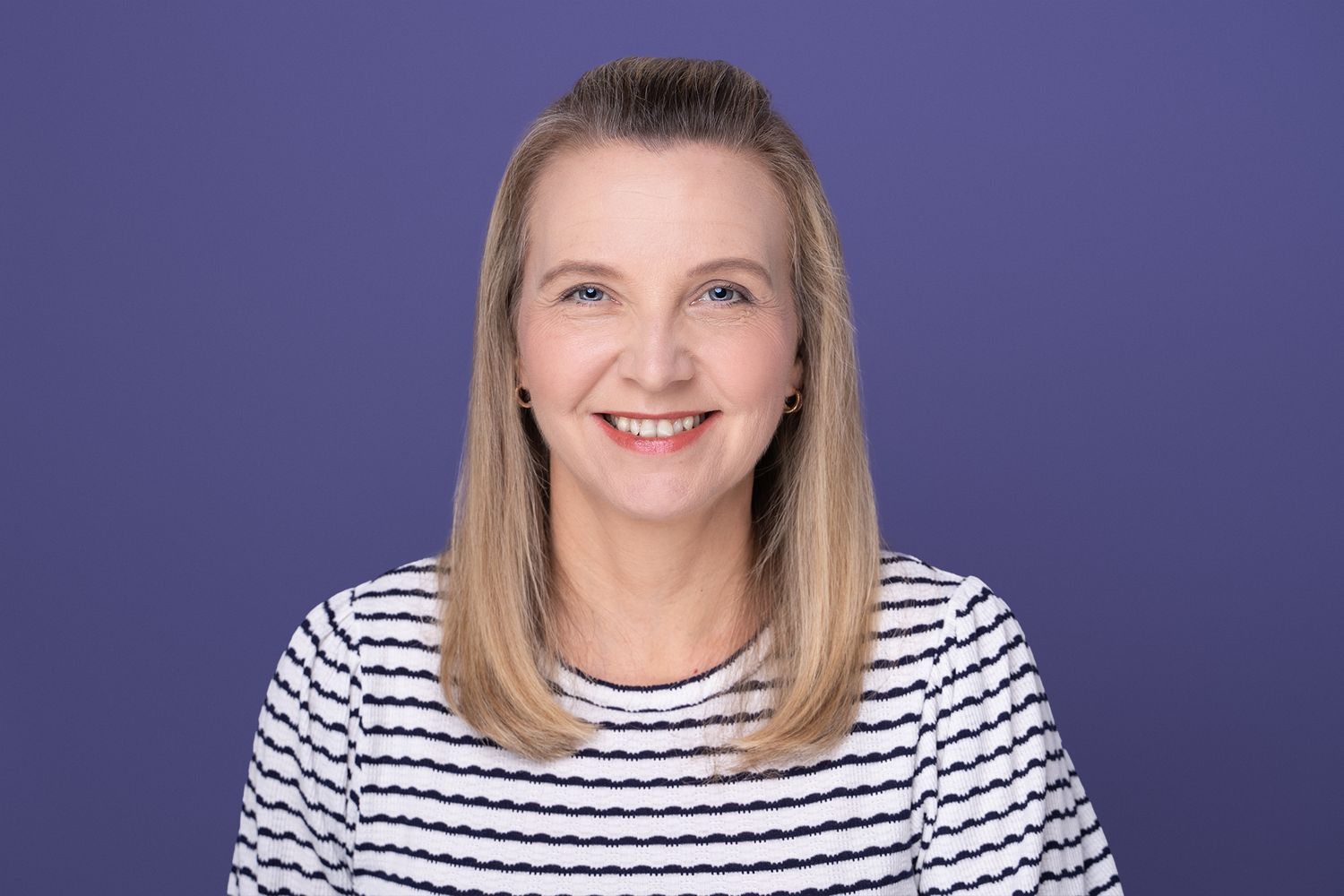 Studio headshot of a Smiling blonde woman, wearing a white striped top, against a purple background