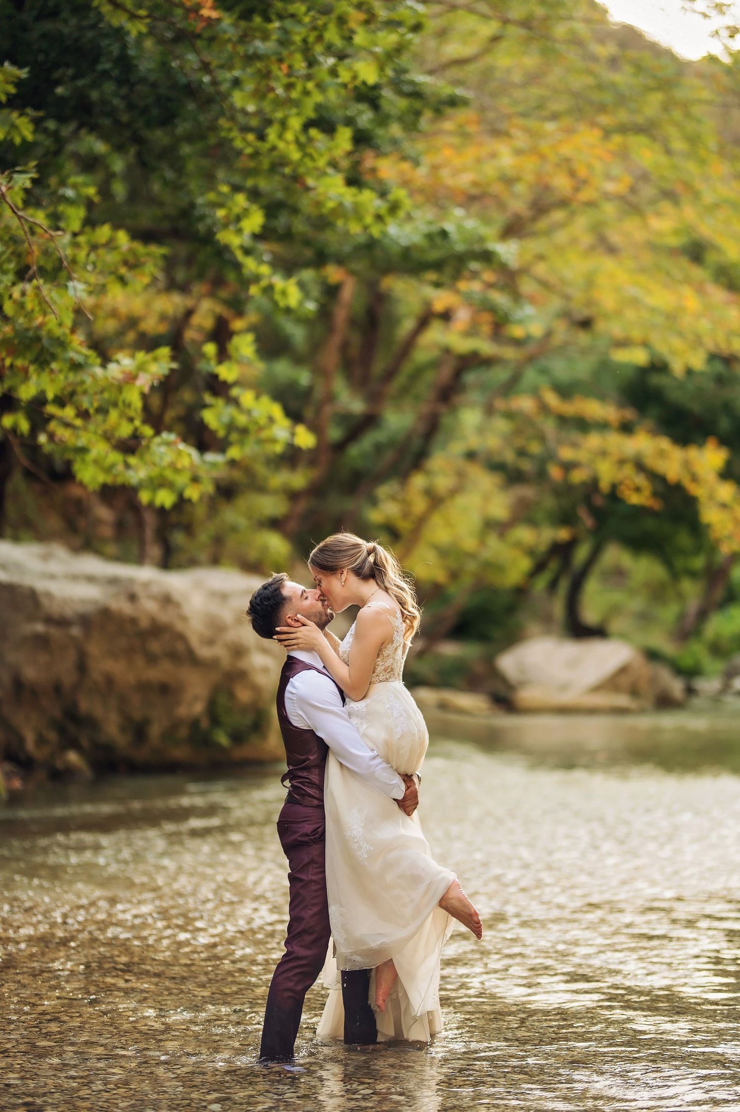 A groom lifts his bride in a shallow river, sharing a romantic kiss surrounded by lush autumn trees.
