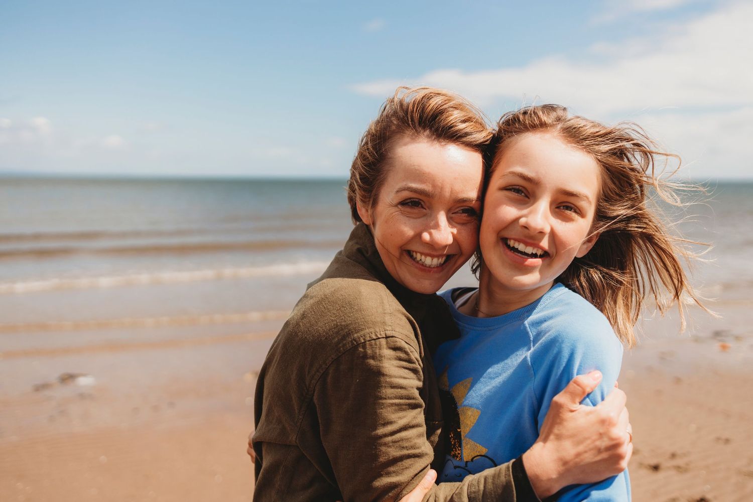 Two women hugging and smiling on a sandy beach with blue sky and ocean in the background.