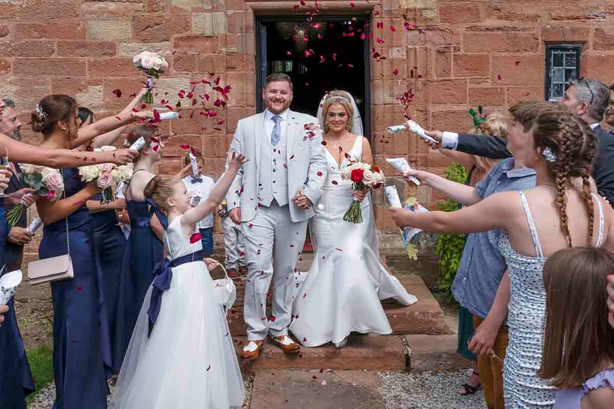 Bride and groom walk through confetti shower outside stone church surrounded by wedding guests in navy and white attire.