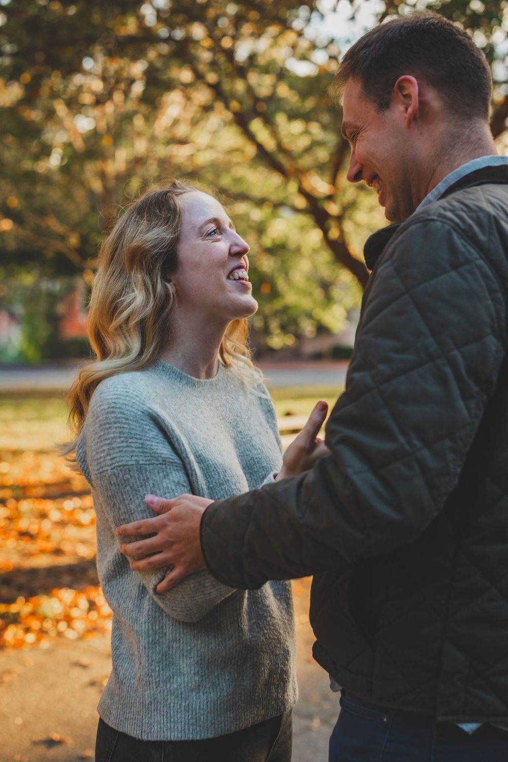 A laughing woman in a gray sweater shares a joyful moment with a man outdoors in an autumn park.