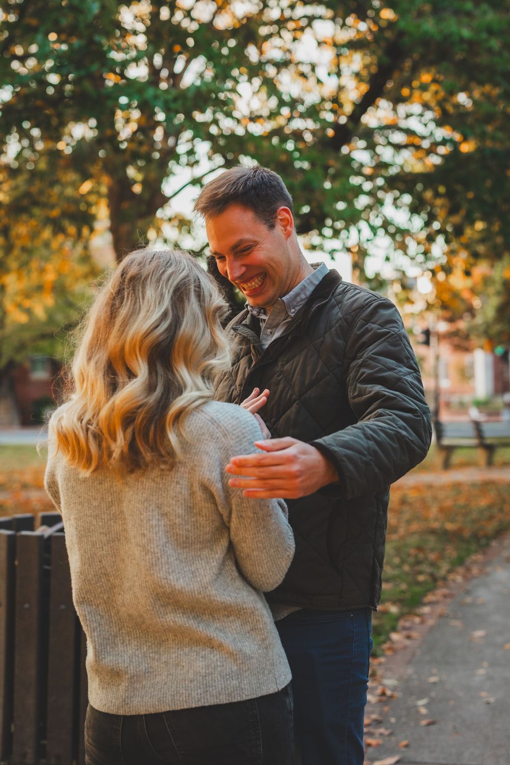A smiling couple embraces in an autumn park, with colorful fall foliage in the background.