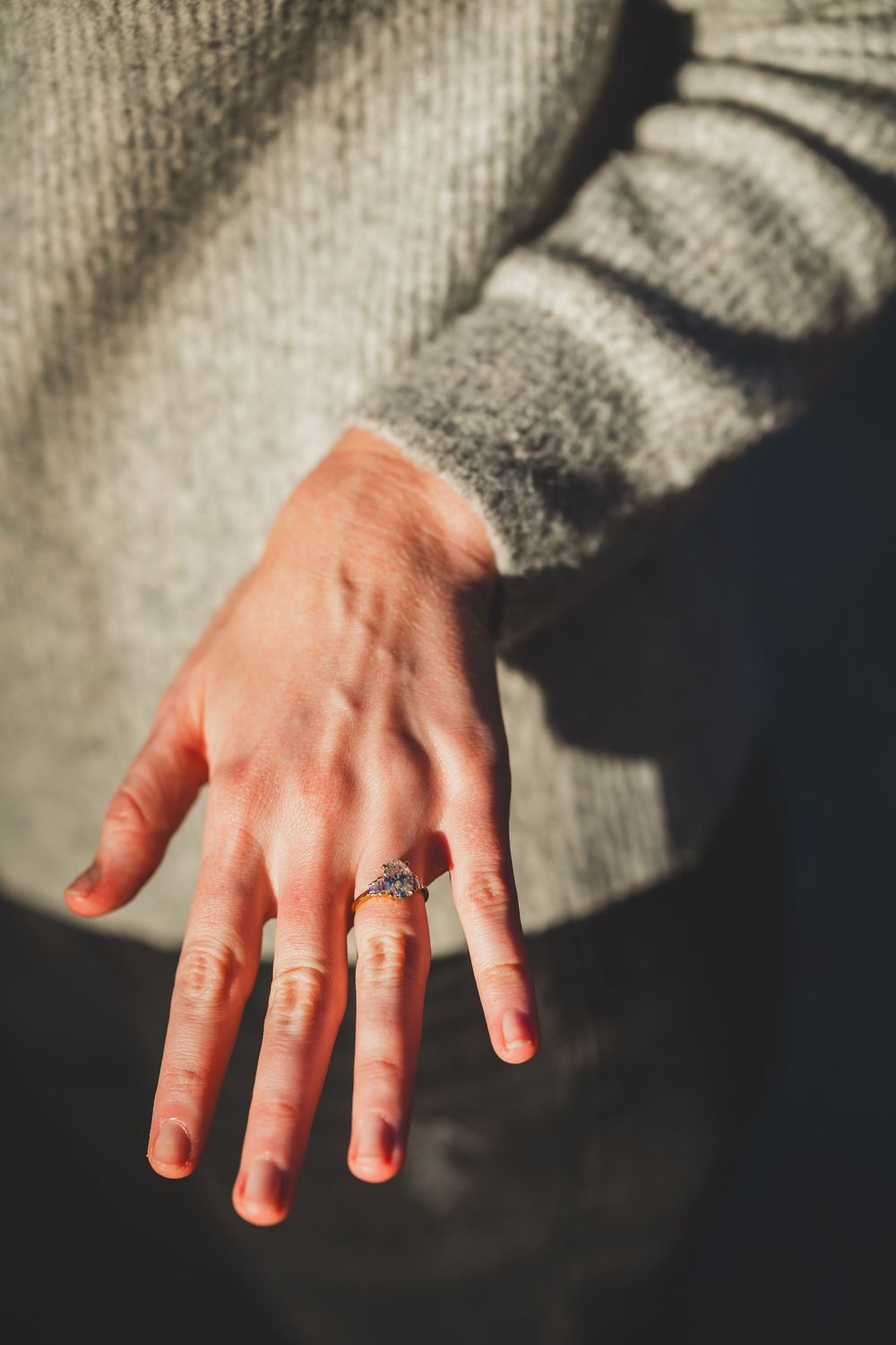 Close-up of a hand wearing a diamond engagement ring, wearing a gray knit sweater sleeve in warm sunlight.