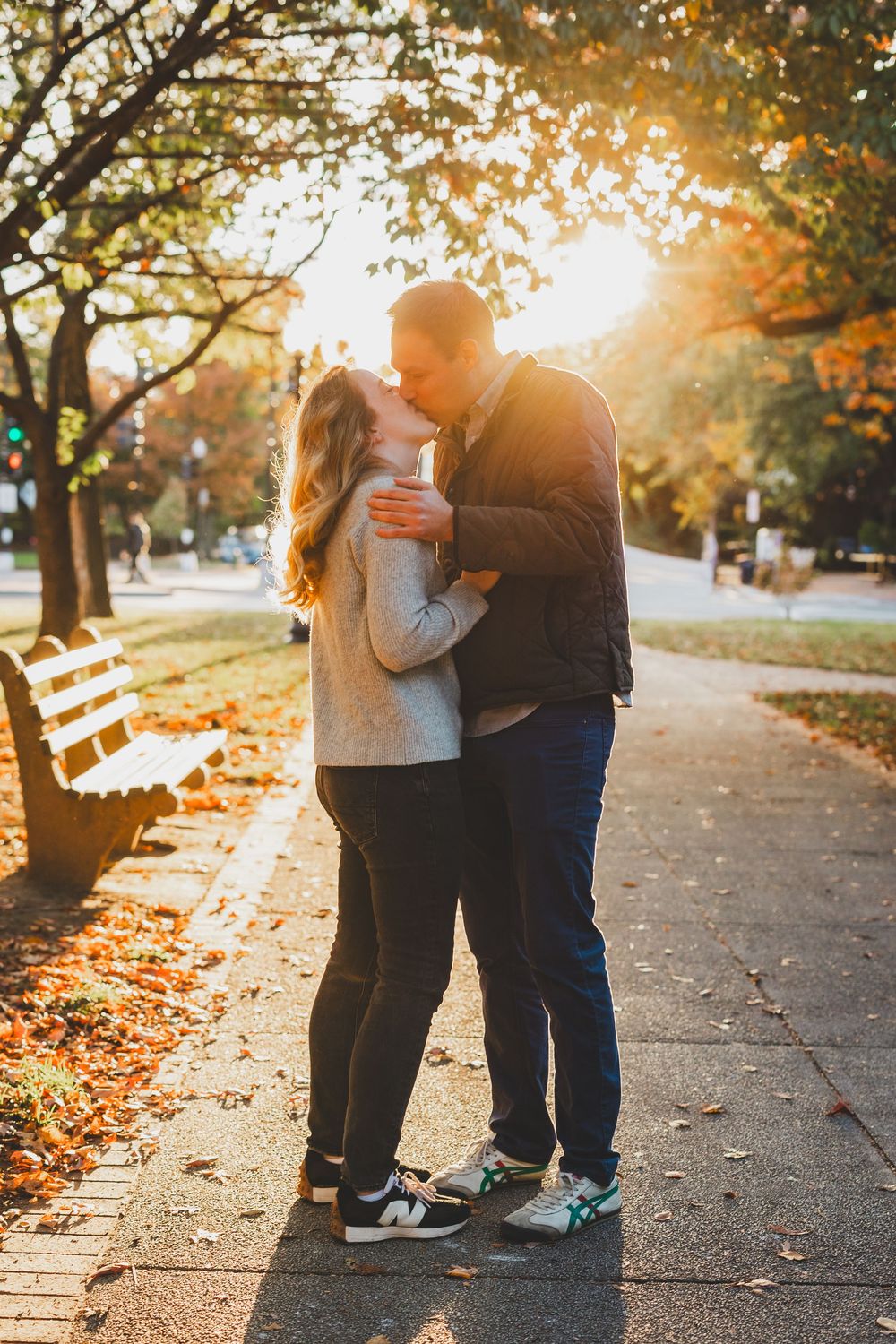 Couple sharing a kiss on a sunlit autumn park path, surrounded by golden fall foliage and a wooden bench.