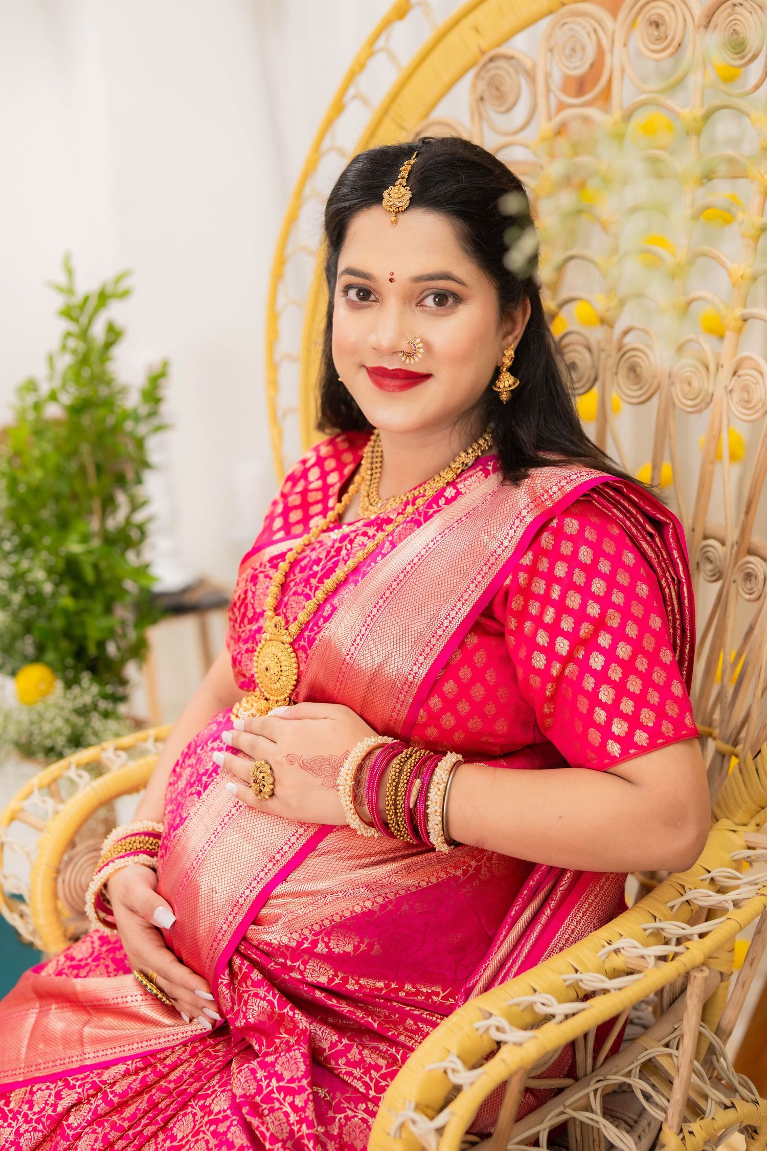 Expecting mother in a pink and gold Banarasi saree seated on a peacock chair with fresh greenery in the background