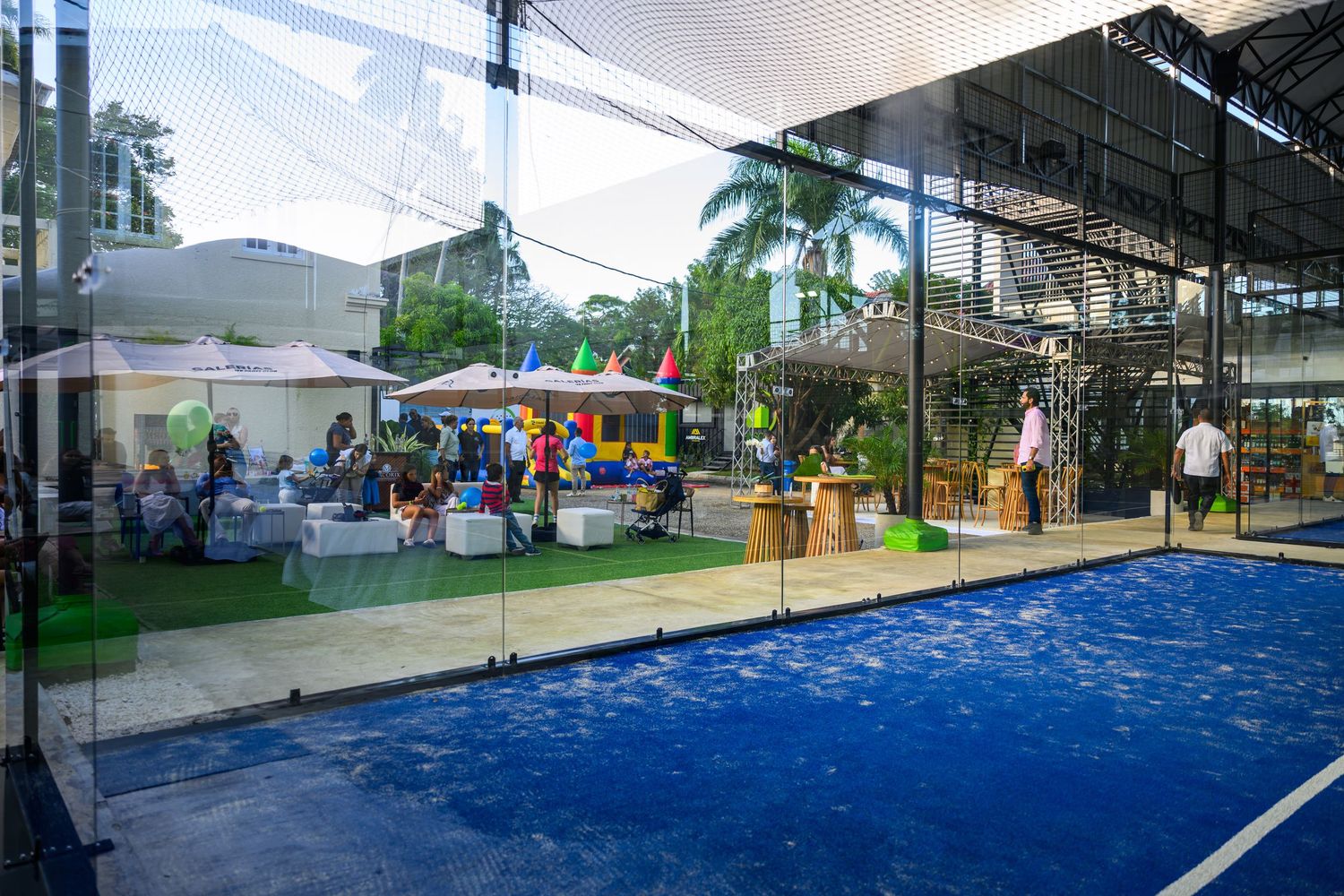 Padel tennis court with blue turf beside an open-air lounge area with colorful umbrellas and seated guests.