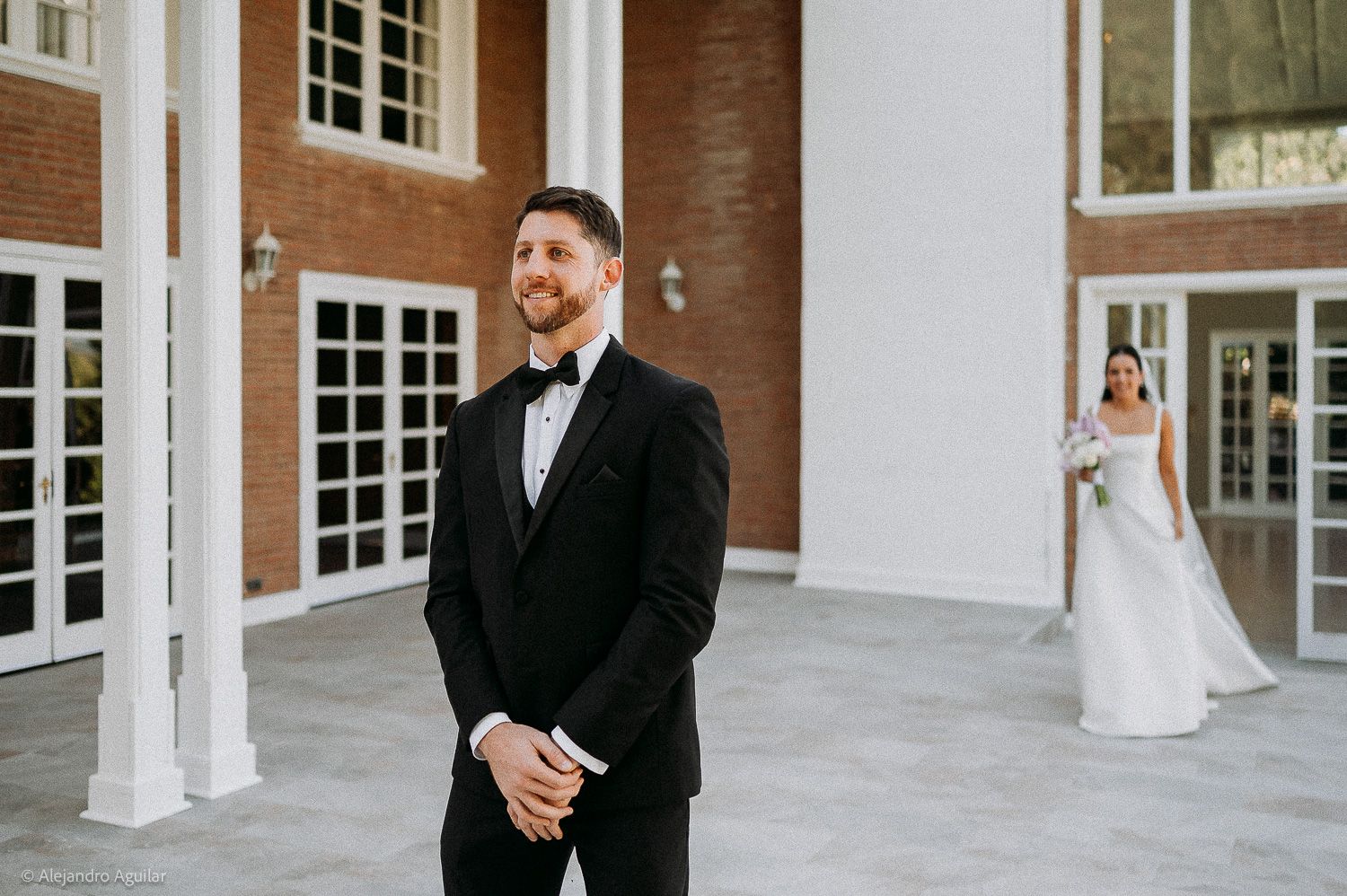 Groom in black tuxedo smiles as bride in white wedding gown approaches from behind at elegant venue.