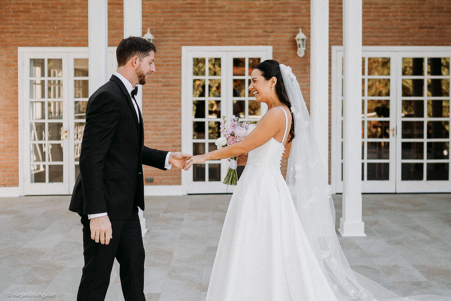 Bride and groom holding hands outdoors at elegant brick venue, bride in white gown with veil, groom in black tuxedo.