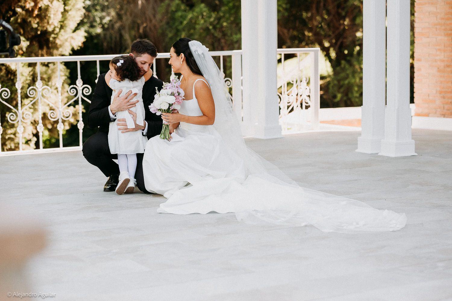 Bride in white gown crouches with groom and flower girl on elegant white outdoor wedding venue terrace.
